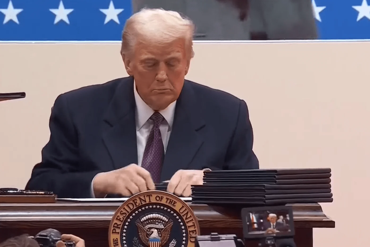 U.S. President Donald Trump signing documents at an official event, with stacks of executive orders or trade agreements in front of him